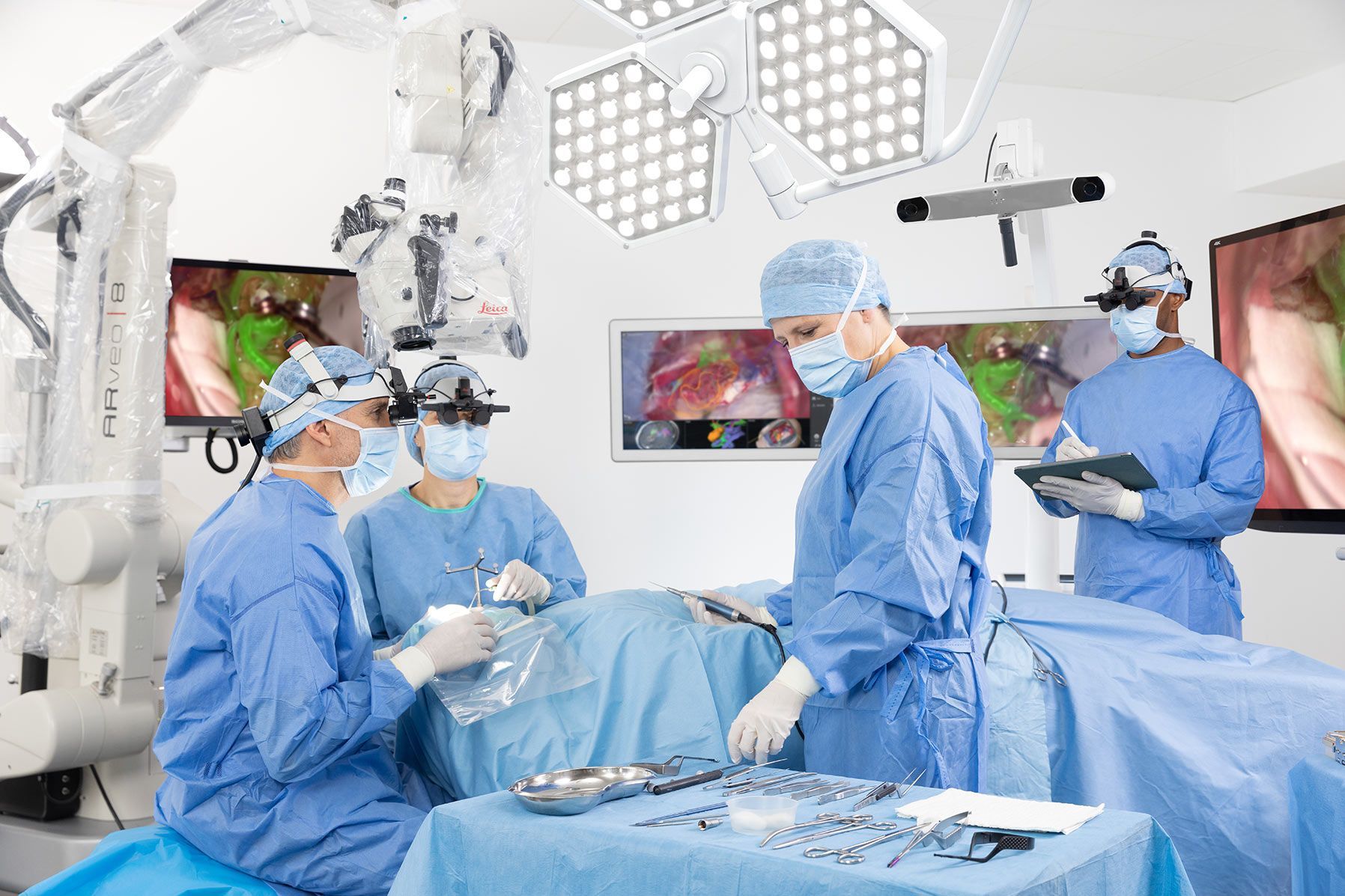 Alt text: A surgeon with his team in the operating room, using the ARveo 8x 3D  digital microscope with GLOW800 for neurovascular surgery and MyVeo headset.