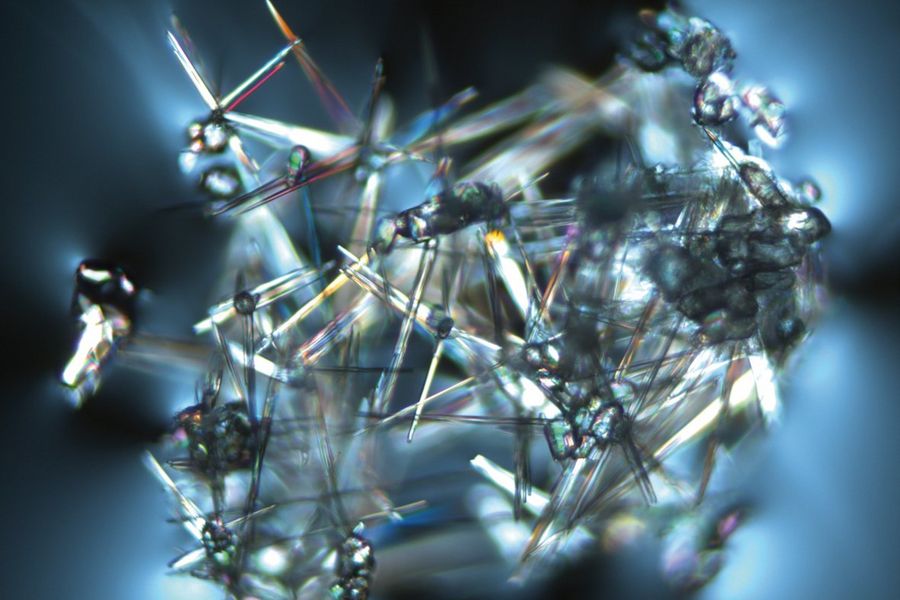 Blue granular tin oxide which remaines next to needle-shaped recrystallisation products. This is an example of a glass inclusion with strong photoelasticity in the surrounding glass. Image was acquired with transmitted light and polarisation contrast using a HC PL Fluotar 10x Pol objective. The image width is 1 mm.